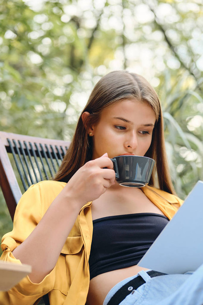 Young attractive thoughtful brown haired woman in yellow shirt and top reading book with coffee on wooden deck chair in city park - Photo, Image