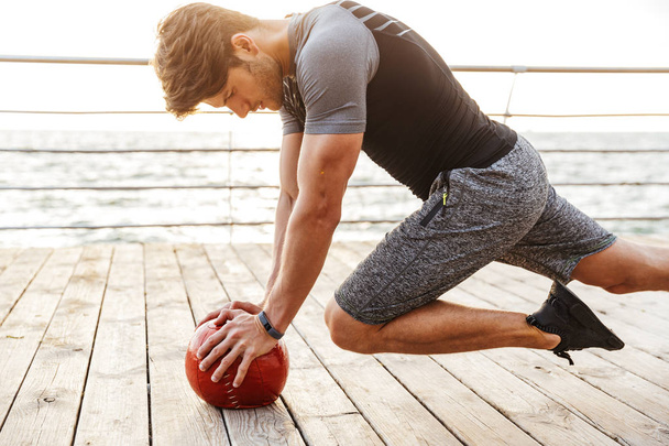 Photo of handsome man in tracksuit doing exercise with red fitness ball while working out on wooden pier at seaside - Photo, image