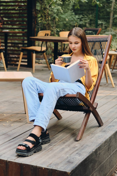 Beautiful brown haired teenage girl in yellow shirt and jeans reading book with cup of coffee on wooden deck chair of cafe in city park - Fotoğraf, Görsel