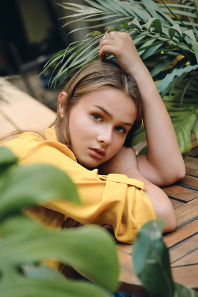 Young serious brown haired woman in yellow shirt lying on hand thoughtfully looking in camera with green leaves around in park - Fotografie, Obrázek