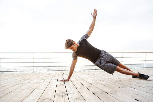 Photo of sporty man in tracksuit raising arm and doing side plank while working out on wooden pier at seaside - Photo, Image