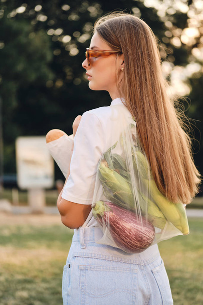 Young attractive brown haired woman standing from back with baguette bread and vegetables in eco bag on shoulder thoughtfully looking aside in city park - Photo, Image