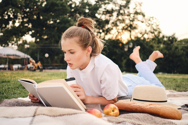 junge attraktive ernsthafte Frau liegt auf Plaid nachdenklich Buch lesen auf Picknick im schönen Stadtpark - Foto, Bild