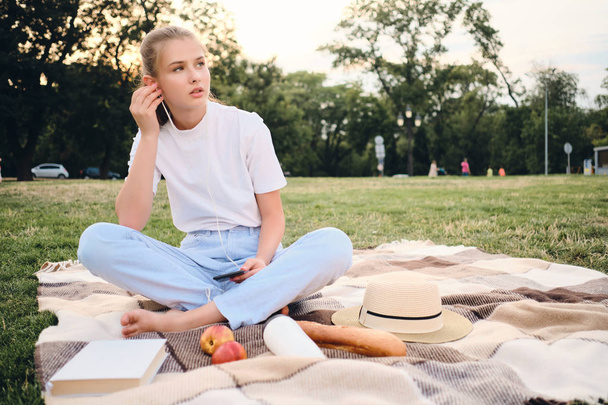 Mooie tiener meisje zittend op Plaid en zorgvuldig luisteren muziek in koptelefoon op picknick in City Park - Foto, afbeelding