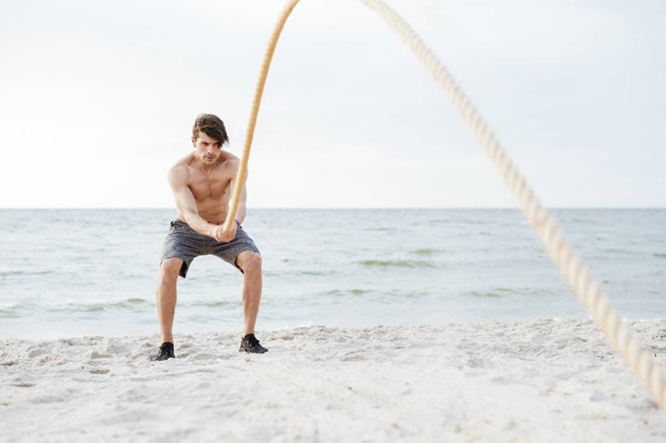Photo of brutal half-naked man working out with fitness rope on beach at seaside - Foto, immagini