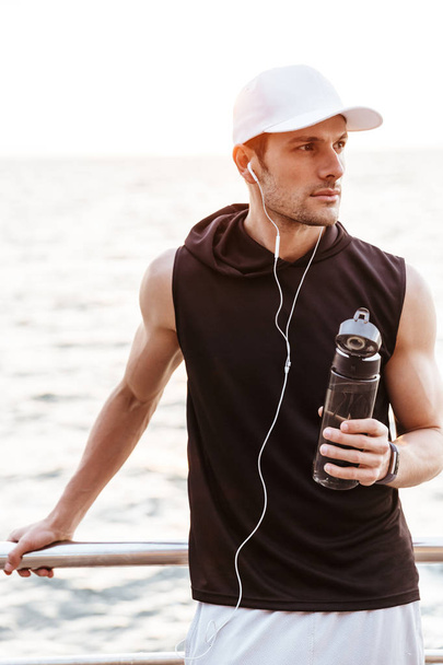 Photo of serious man in white cap using earphones and drinking water while walking on pier at seaside - Fotografie, Obrázek