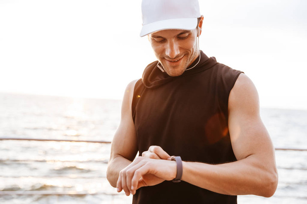 Photo of smiling man in white cap listening to music with earphones and using smart watch while walking at seaside - Foto, immagini