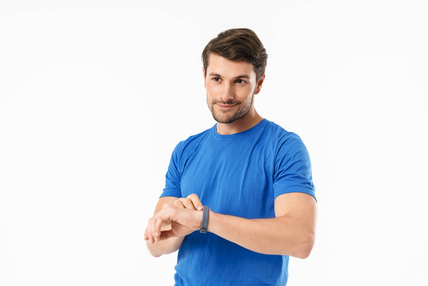 Happy smiling handsome young sports fitness man standing isolated over white wall background using watch clock. - Foto, imagen