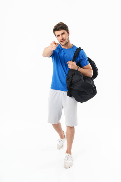 Serious handsome young sports fitness man standing isolated over white wall background holding bag pointing to you. - Φωτογραφία, εικόνα