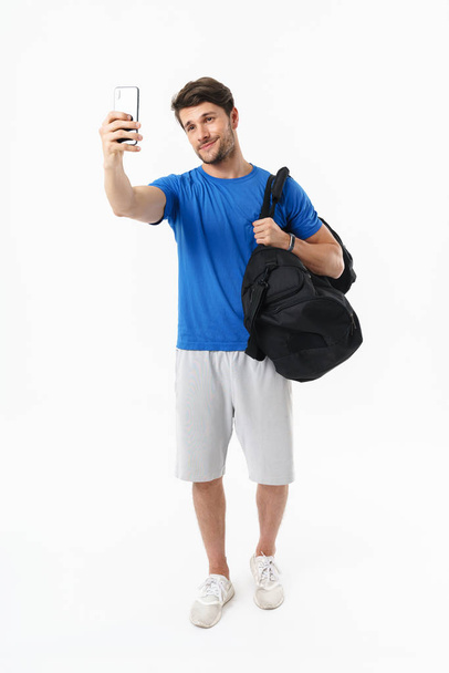 Pleased smiling handsome young sports fitness man standing isolated over white wall background holding bag make selfie by mobile phone. - Foto, immagini