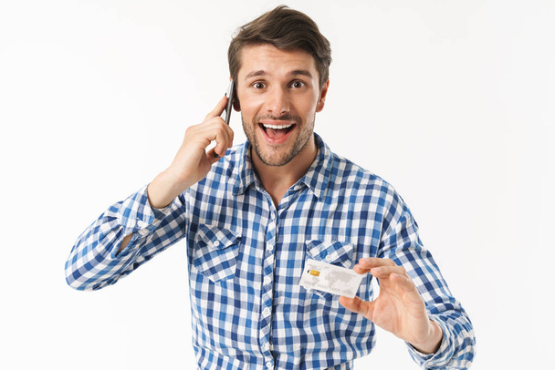 Excited cheerful handsome young man standing isolated over white wall background dressed in casual shirt talking by mobile phone holding credit card. - Foto, immagini
