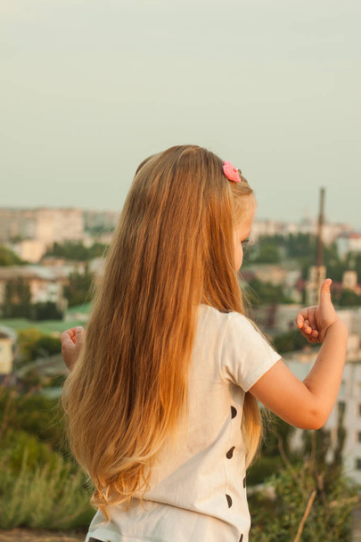Μικρό κορίτσι με μακριά ξανθά μαλλιά σε ένα λευκό T-shirt σε φόντο φύσης. - Φωτογραφία, εικόνα