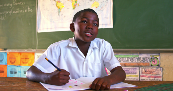 Front view close up of a young African schoolboy sitting at a desk looking up while writing in his note book and listening attentively during a lesson in a township elementary school classroom 4k - Video, Çekim