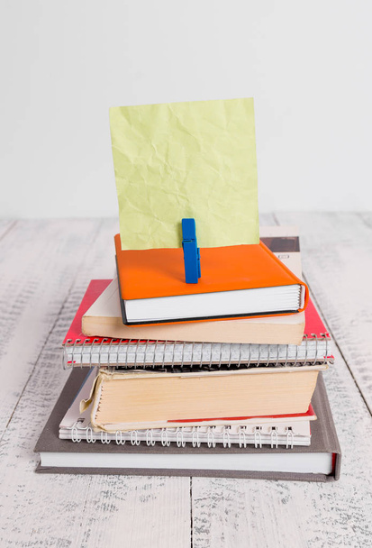 Square shaped paper above a pile of notebooks fixed by a blue clothespin. Different books stacked under an empty reminder colorful note. White wooden floor and white background. - Photo, image