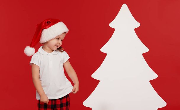 Happy little child girl in Santa hat at the Christmas time. Isolated red background. Empty place for an inscription in the form of a Christmas tree - Fotoğraf, Görsel