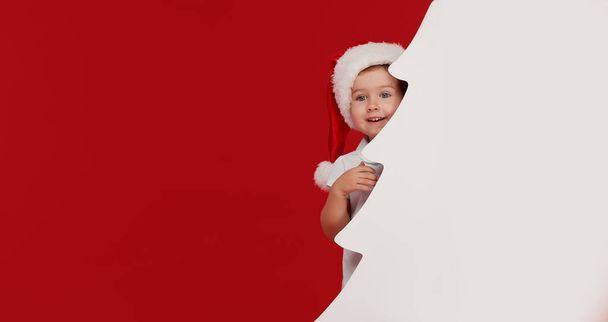 Happy little girl child in Santa hat peeking from behind blank sign banner in the shape of Christmas trees. Isolated on red background. Space for Your Text. Sale, holidays, christmas, new year concept - Fotoğraf, Görsel