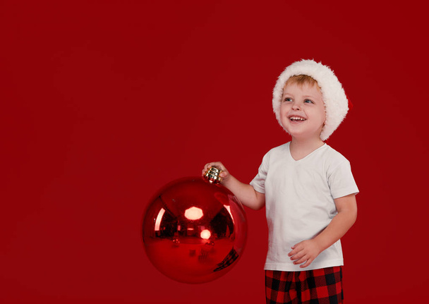 Little kid boy in santa claus hat holds big christmas ball on red isolated background. Point finger left, blank space for text. - Photo, Image