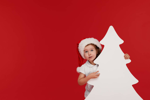 Happy little girl child in Santa hat peeking from behind blank sign banner in the shape of Christmas trees. Isolated on red background. Space for Your Text. Sale, holidays, christmas, new year concept - Photo, image
