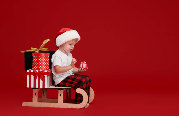 Adorable little child wearing red pajamas and santa claus hat, sitting in a wooden Christmas snow sleigh. Bears children holiday gifts in boxes - Foto, Imagem