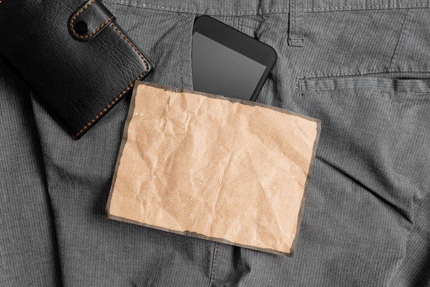 Smartphone-apparaat in de broek voorvak met een portemonnee en bruine notitie papier. Moderne gadget is geplaatst in man broek in de buurt van notatie blad plus portemonnee - Foto, afbeelding