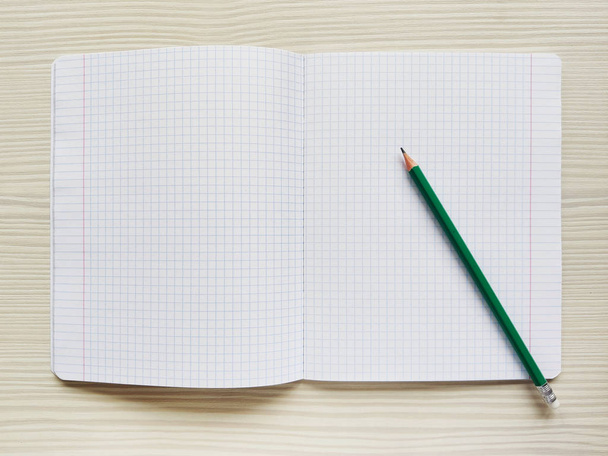 White school notebook in a cage is on a wooden table. Green pencil rests on a checkered exercise book without notes. Empty notepad is on a desk. Square texture - Φωτογραφία, εικόνα