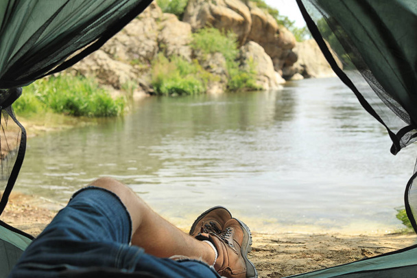 Young man resting in camping tent on riverbank, view from inside - Foto, Imagen
