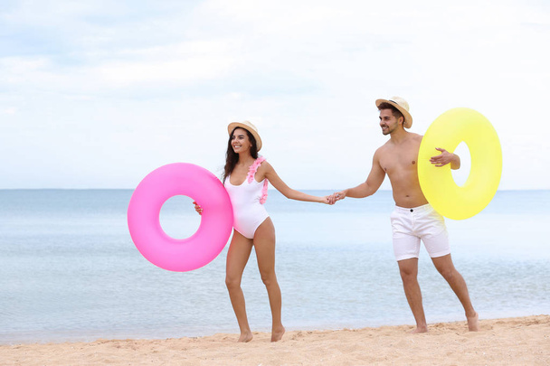 Happy young couple having fun with inflatable rings on beach near sea - Foto, afbeelding