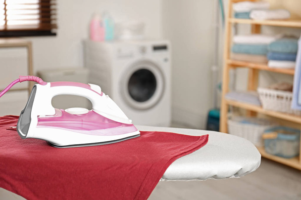 Modern electric iron and clean t-shirt on board in laundry room. Space for text - Zdjęcie, obraz