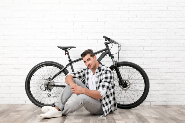 Handsome young man with modern bicycle near white brick wall indoors - Foto, afbeelding