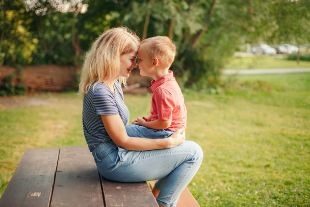 Młoda Blondynka kaukaski matka i chłopiec maluch syn siedzi twarzą w twarz na drewnianej ławce na zewnątrz w letni dzień i rozmawia ze sobą. Szczęśliwego życia rodzinnego z dzieciństwa.  - Zdjęcie, obraz