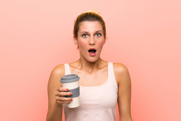 Νεαρή ξανθιά γυναίκα κρατώντας ένα πάρει μακριά καφέ με έκπληξη και σοκαρισμένος έκφραση του προσώπου - Φωτογραφία, εικόνα