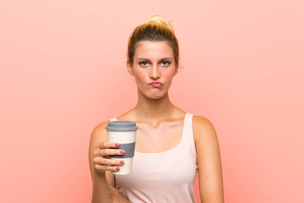 Νεαρή ξανθιά γυναίκα κρατώντας ένα πάρει μακριά καφέ με θλιβερή έκφραση - Φωτογραφία, εικόνα