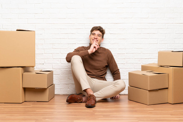 Handsome young man moving in new home among boxes doing silence gesture - Foto, afbeelding