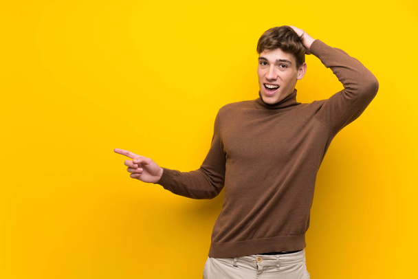 Красивый молодой человек на изолированном желтом фоне удивлен и указывая пальцем в сторону
 - Фото, изображение