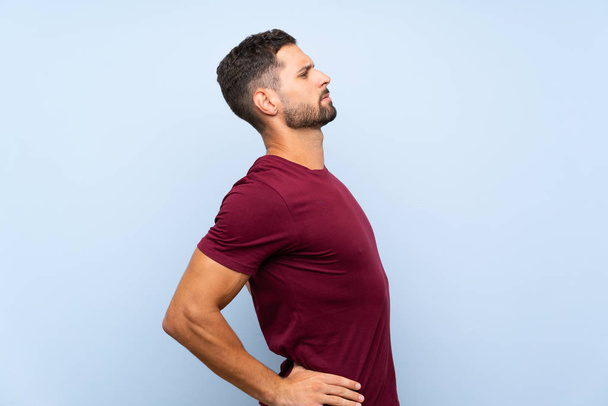 Knappe man over geïsoleerde blauwe achtergrond die lijden aan rugpijn voor het hebben van een inspanning - Foto, afbeelding