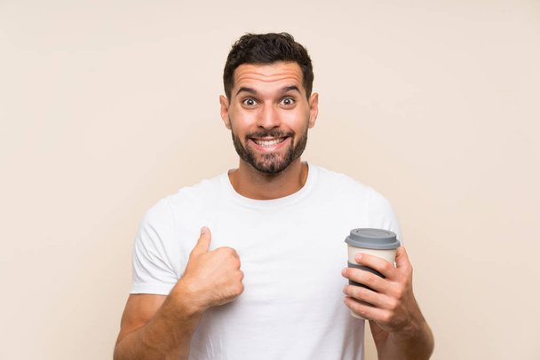 junger Mann mit Bart, der einen Take-away-Kaffee vor isoliertem blauen Hintergrund mit überraschendem Gesichtsausdruck hält - Foto, Bild