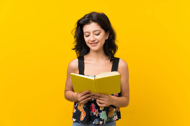 Mujer joven sobre fondo amarillo aislado sosteniendo y leyendo un libro
 - Foto, Imagen