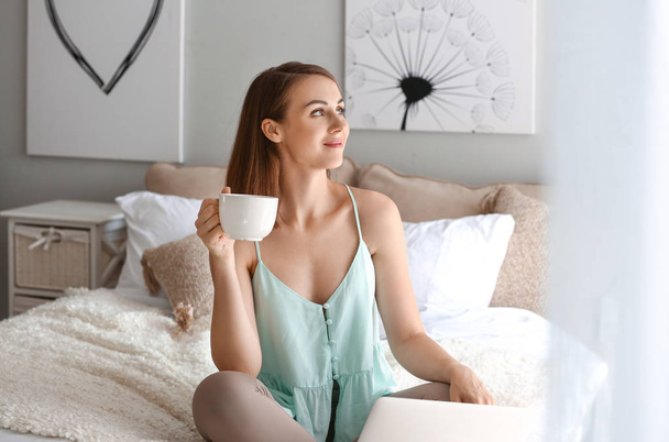 Όμορφη νεαρή γυναίκα με φορητό υπολογιστή πίνοντας τσάι στο σπίτι - Φωτογραφία, εικόνα