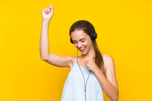 Jonge vrouw luisteren muziek over geïsoleerde gele muur vieren een overwinning - Foto, afbeelding