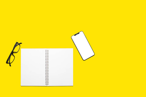 黄色の分離背景にコピースペースを持つ空白のノートブック用紙とスマートフォンの画面のトップビュー - 写真・画像