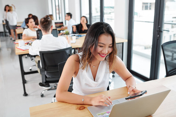 Uśmiechnięta Azjatycka bizneswoman siedzi w jej miejscu pracy za pomocą telefonu komórkowego i wpisując na laptopie komputer z ludźmi biznesu mają spotkanie w tle - Zdjęcie, obraz