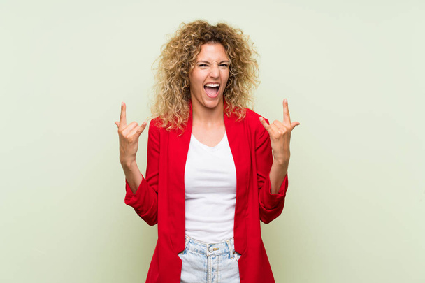 Young blonde woman with curly hair over isolated green background making rock gesture - Fotoğraf, Görsel