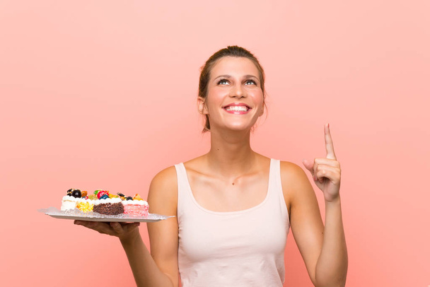 Young blonde woman holding lots of different mini cakes pointing up a great idea - Foto, Bild