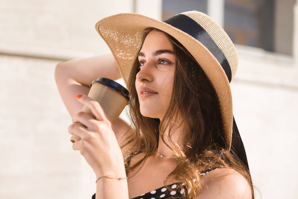 Pretty girl in a black dress, hat with wide flaps and sun glasses holds disposable paper cup of hot coffee enjoying walk down the European town during sunny summer day. - Foto, Imagem