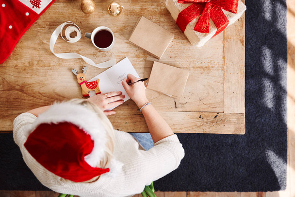 Kopfschuss auf Frau, die in Weihnachtskarte schreibt und Geschenk mit Weihnachtsmütze einwickelt - Foto, Bild