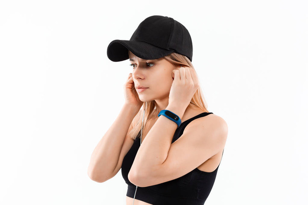 スマートウォッチ付きの黒いスポーツウェアを着た若いスポーティーなブロンドの女性は、白い背景の上に立ってワークアウト中に音楽を聴きます. - 写真・画像