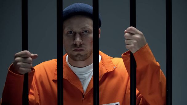 Dangerous imprisoned criminal with scar on face holding bars looking at camera - Foto, Imagem