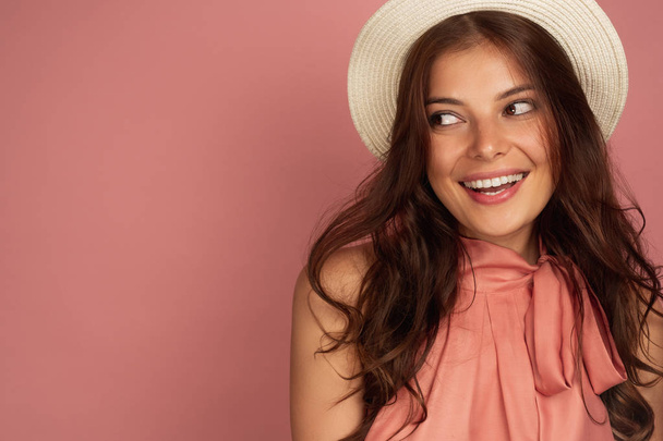The brunette is standing on a pink background in a straw hat and smiling cheerfully looks to the side. - Photo, Image