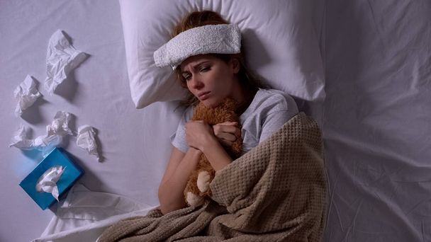 Lonely sick woman hugging teddy-bear lying in bed with compress and tissues - Photo, Image
