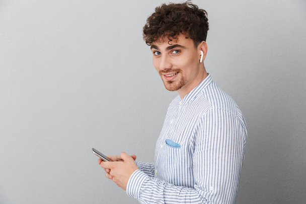 Image of cheerful young man with earpods smiling at camera and holding cellphone - Fotografie, Obrázek